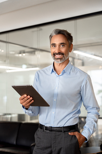 Vertical portrait of smiling Hispanic mature adult professional business man, happy 40s 50s businessman CEO holding digital tablet using fintech tab application standing in office, looking at camera.