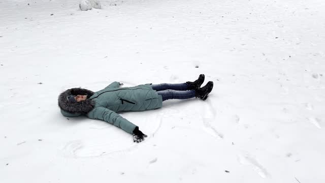 Happy woman lying on snowy lawn at forest and making angel. Young girl playing in snow and enjoying wintertime. Cheerful lady having fun outdoor. Concept of a winter vacation. Close up Slow motion