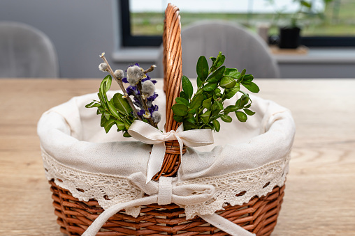 An empty wicker Easter basket, decorated with boxwood and catkins, standing on a table.