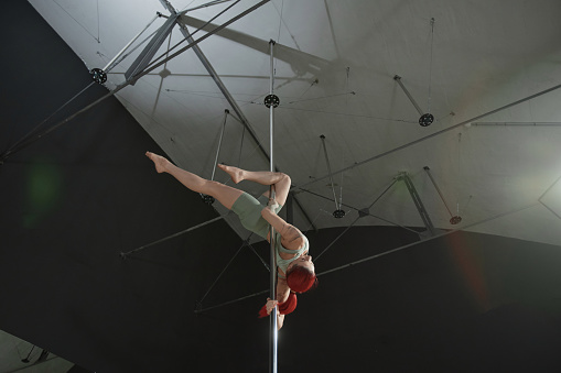 Low angle view of unrecognizable red-haired woman training pole dance elements hanging upside down under studios ceiling