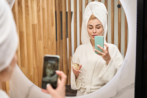 Blonde woman in the bathroom takes a selfie in front of the mirror, she is in a fluffy bathrobe