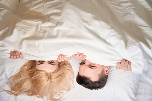 Man and a woman sit comfortably under a blanket on a large bed, a couples room for newlyweds
