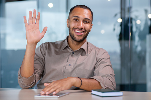 Friendly Arab manager in the office talking on a video call with clients, working on a laptop, greeting, waving at the camera, webcam view.