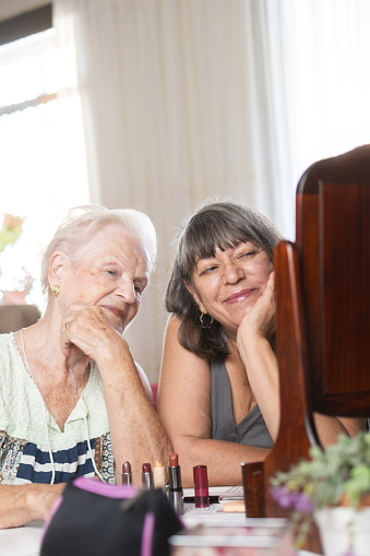 mother and daughter making up together and smiling in front of the vanity mirror.love and bonding in family time.two generations of women.simple living and well-being at home.vertical image.