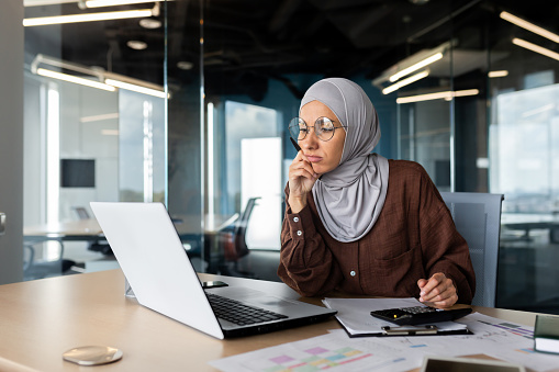Tired young Muslim woman in hijab is sitting in the office at the desk and looking bored at the monitor screen.