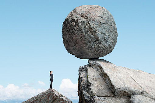 A businesswoman stands and looks up a a large round boulder that is precariously balanced on the rocky promontory above her.