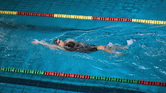 Female athlete in action, performing the backstroke swim technique in the indoor lap pool. Competitive back crawl stroke concept.