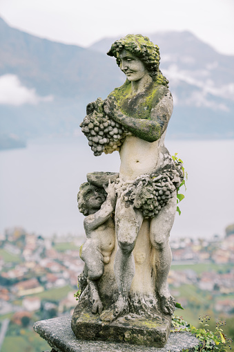 Antique statue of the ancient Greek god Dionysus with a bunch of grapes with a small faun with a bowl in the garden. High quality photo