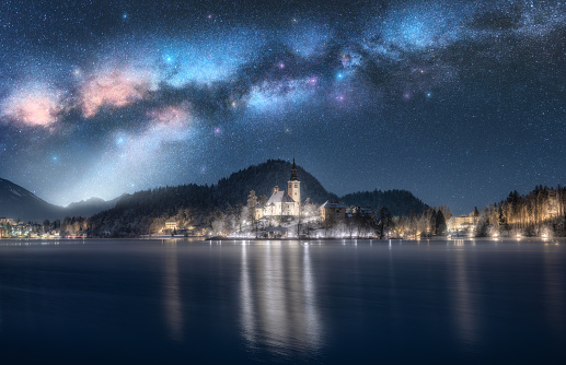 Milky Way over the church on the snowy island on the Bled Lake, Slovenia at winter starry night. Landscape with chapel, illumination, sky with stars, mountains, street lights, reflection in water