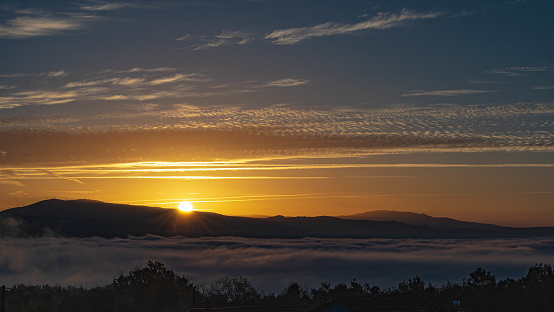 The sun begins to rise, illuminating the sky with high clouds and leaving the valley in shadows covered with fog