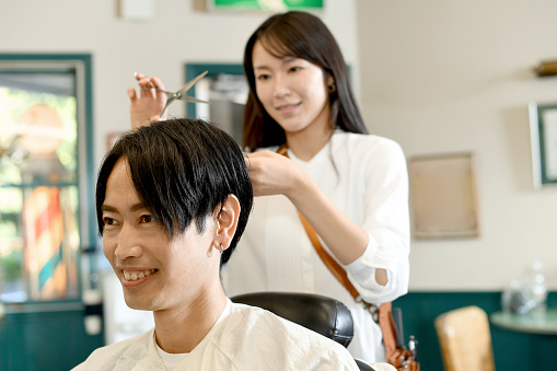 Smiling Asian male customer having his hair cut with scissors by a young Asian female hairdresser