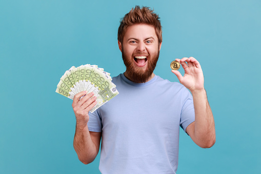 Portrait of positive optimnistic handsome bearded man showing euro banknotes and bitcoin, digital money, electronic commerce, expressing excitement. Indoor studio shot isolated on blue background.