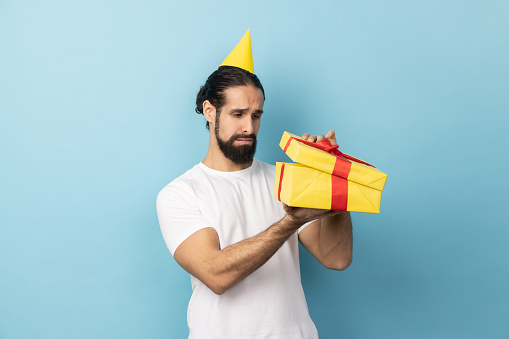 Portrait of unhappy man with beard wearing white T-shirt and party cone looking inside present box and expressing sadness, hoped to get another gift. Indoor studio shot isolated on blue background.