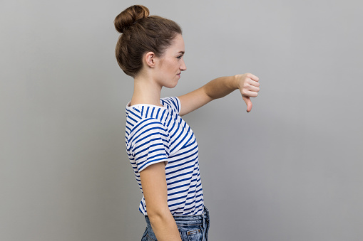 Side view of unhappy unsatisfied young adult woman wearing striped T-shirt showing thumb down, dislike gesture, negative emotions, bad rating. Indoor studio shot isolated on gray background.