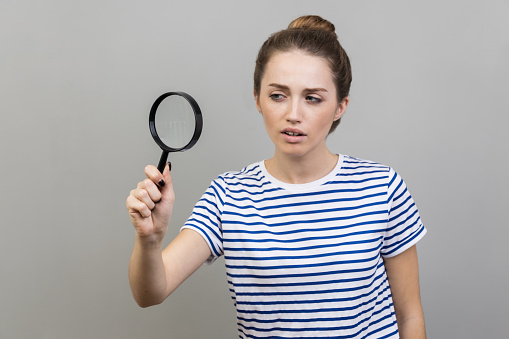 Portrait of woman wearing striped T-shirt looking through magnifying glass, finding out something, magnifies small details to see, inspecting. Indoor studio shot isolated on gray background.