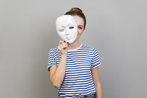 Portrait of woman wearing striped T-shirt removing white mask from face showing his smiling expression, good mood, pretending to be another person. Indoor studio shot isolated on gray background.