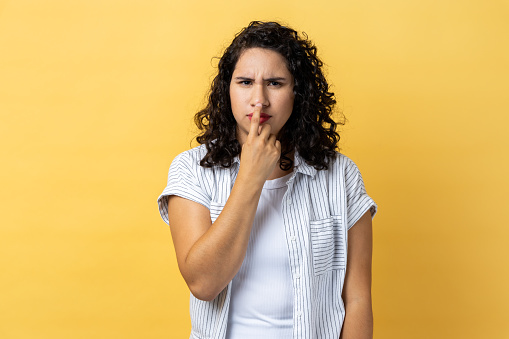 Portrait of woman with dark wavy hair looking with incredulous suspicious gaze and touching nose, gesturing you are liar, suspecting falsehood. Indoor studio shot isolated on yellow background.
