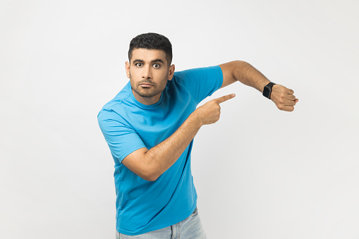 Time to go, hurry up. Portrait of unshaven man in blue T- shirt standing pointing at wrist, makes time gesture, shows we should do everything quickly. Indoor studio shot isolated on gray background.