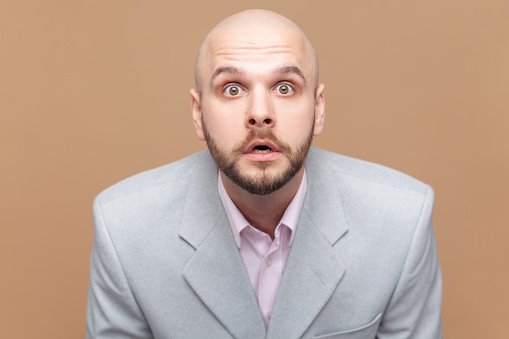Portrait of bald bearded man looks with bugged eyes at camera, being surprised to see big sales, being afraid of something, wearing gray jacket. Indoor studio shot isolated on brown background.