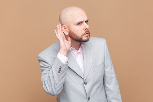 Portrait of bald man overhearing, listening intently to secret information, private talk, holding hand near ear to hear better, wearing gray jacket. Indoor studio shot isolated on brown background.
