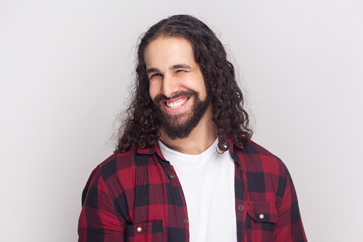 Bearded man with long curly hair in checkered red shirt winking while having good mood smiling, blinking eye showing his sympathy or positiveness. Indoor studio shot isolated on gray background.