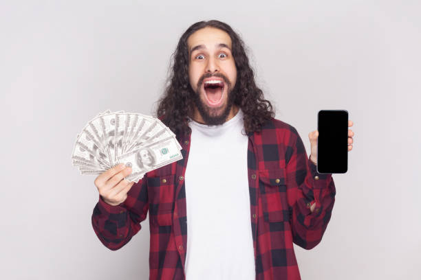 istock Amazed excited bearded man holding dollar banknotes and cell phone with mockup blank display. 1966121941