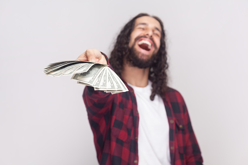 Portrait of joyful cheerful laughing bearded man with long curly hair in checkered red shirt giving you you winning, holding out big fan of money. Indoor studio shot isolated on gray background.