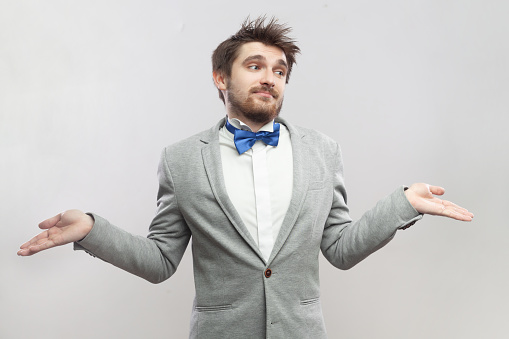 Portrait of uncertain handsome bearded man spreads hands, shrugging shoulders, doesn't know how to find solution, wearing grey suit and blue bow tie. Indoor studio shot isolated on gray background.