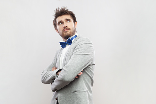 Portrait of confident pensive thoughtful bearded man standing folded hands, looking away, planing and pondering, wearing grey suit and blue bow tie. Indoor studio shot isolated on gray background.