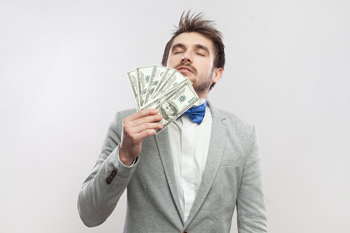Portrait of rich delighted proud bearded man standing smelling dollar banknotes, being satisfied of big salary, wearing grey suit and blue bow tie. Indoor studio shot isolated on gray background.