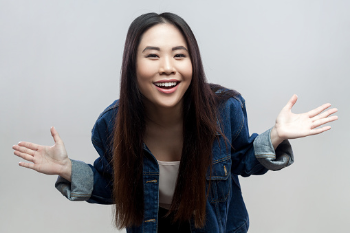 Portrait of positive brunette woman in blue denim jacket standing standing with spread hands, looking at camera and laughing. Indoor studio shot isolated on gray background.
