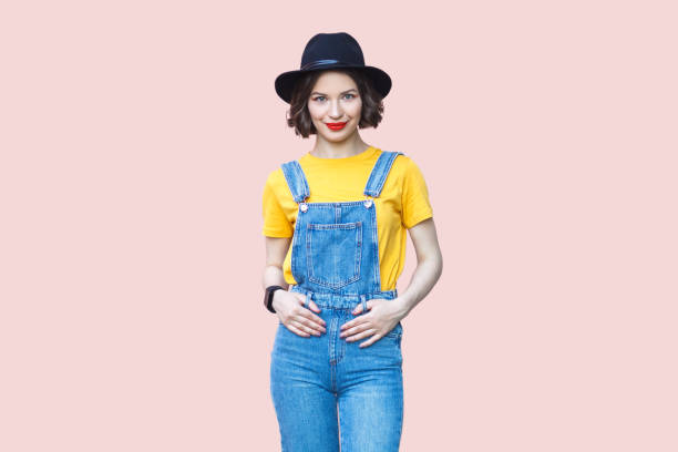 Joyful hipster woman in blue denim overalls looking at camera with happy expression.
