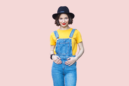 Portrait of smiling joyful hipster woman in blue denim overalls, yellow T-shirt and black hat looking at camera with happy expression. Indoor studio shot isolated on light pink background.