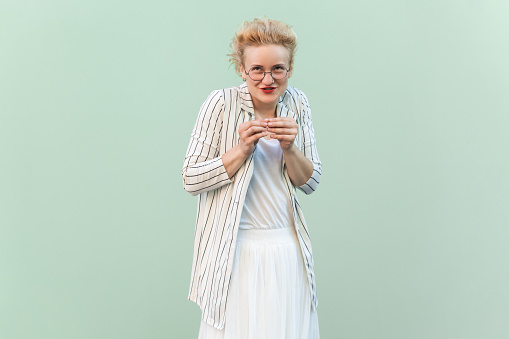 Portrait of cunning young adult blonde woman wearing striped shirt and skirt, planning devil prank, looking at camera. Indoor studio shot isolated on light green background.