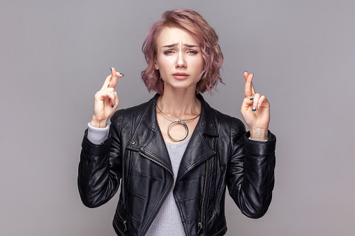 Portrait of winsome woman with short hairstyle standing with crossed fingers, making for good luck, looking at camera, wearing black leather jacket. Indoor studio shot isolated on grey background.