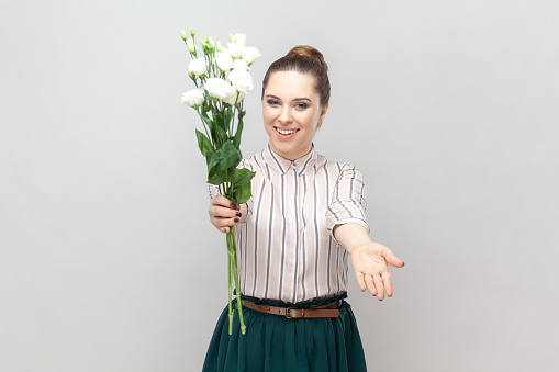 Portrait of cheerful pretty attractive woman wearing striped shirt and green skirt holding bouquet of white flowers, spreading hand, giving it for you. Indoor studio shot isolated on gray background.