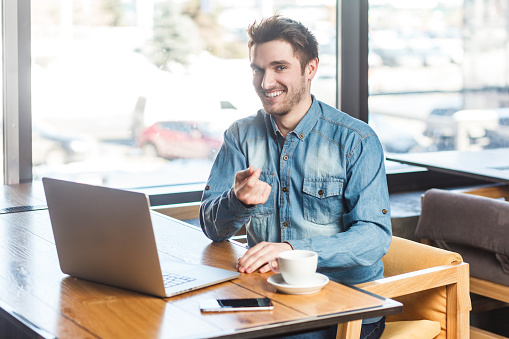Portrait of delighted satisfied smiling bearded young man freelancer in blue jeans shirt working on laptop, pointing finger to camera. Indoor shot near big window, cafe background.