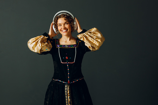 Portrait of a smiling renaissance woman with headphones and touching head and dressed in a medieval dress listens music