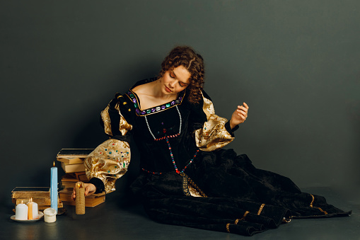 Young adult woman dressed in a medieval dress sitting near ancient books and candle light