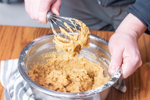 A silver mixing bowl sits on a wooden kitchen table, a towel underneath the bowl to keep it steady. A person is holding the bowl with their left hand and a metal whisk in their right. The whisk is covered in tan colored cookie dough.