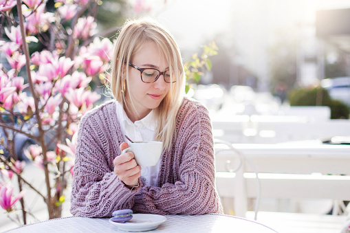 Young woman is drinking coffee in cafe on spring city streets. Blooming bushes of flowers are pink and purple. Attractive girl is enjoying of magnolia garden at sunset outside.