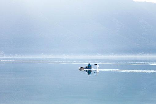 Blue background. Kayaking in foggy morning lake. Boat with aged retired sportsman on sea. Old man has active lifestyle in seniority. Amazing nature landscape with mountains. Copy space.