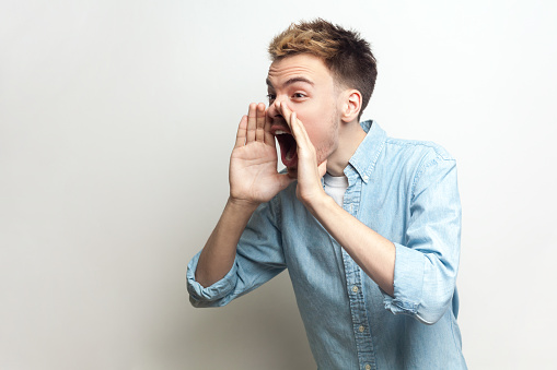 Portrait of angry aggressive attractive man wearing denim shirt standing with hands near his mouth, screaming loud, announcing bad news. Indoor studio shot isolated on gray background.