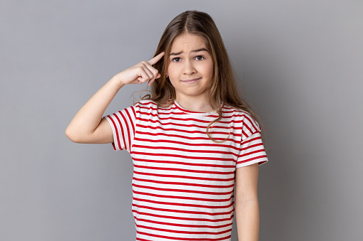 Are you kidding me? Portrait of little girl wearing striped T-shirt holding finger near head temple and gesturing, looking displeased with crazy idea. Indoor studio shot isolated on gray background.