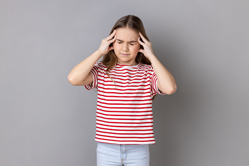 Little girl wearing striped T-shirt frowning, clasping sore head, suffering intense headache, having unbearable migraine, fever and flu symptoms. Indoor studio shot isolated on gray background.