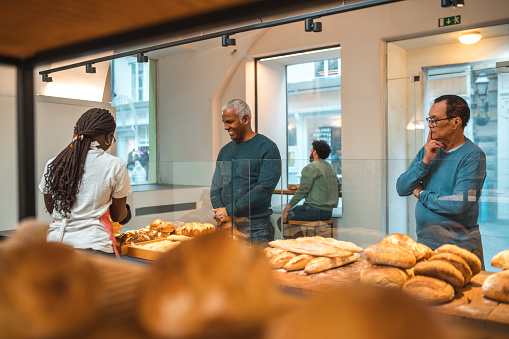 Diverse male and female customers standing in line in a small artisan bakery. Buying freshly baked goods. Black female shop assistant handing them their orders.