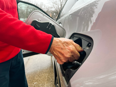 Partial view of a senior man at a gas station pumping gas into his car. He is dressed casually in red and navy blue gear. His hand is tightening the knob after pumping gas. The hand is that of an older man with arthritis.