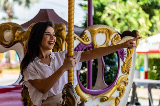 The asian young woman happily pointing her finger to the rignt with smiling and sitting in the carousel of the amusement park