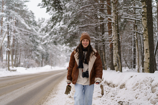 Portrait of smiling stylish asian female blogger traveler dressed in warm sheepskin coat walking towards camera along snowy road and winter forest, happy woman enjoying hiking and frosty nature around