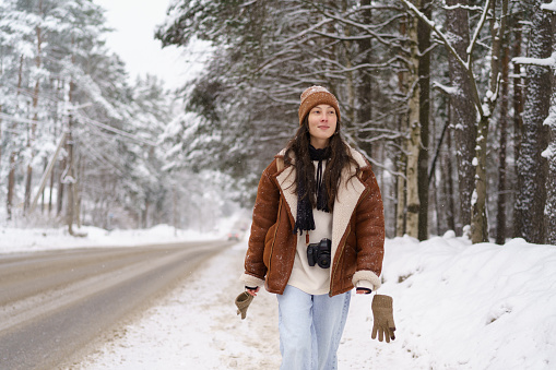 Stylish young asian woman influencer or famous travel blogger with photo camera around neck strolling along snowy forest road on cold winter day while traveling travel out of town in wintertime